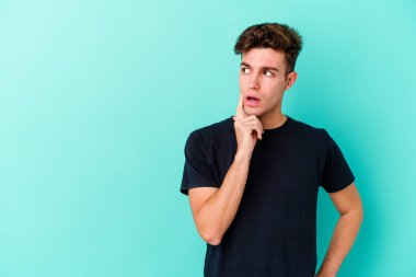 Mavi arka planda izole edilmiş genç beyaz adam şüpheli ve şüpheci bir ifadeyle yana bakıyor..