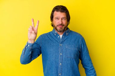 Sarı arka planda izole edilmiş orta yaşlı beyaz bir adam zafer işareti gösteriyor ve gülümsüyor..