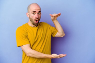 Mor arka planda izole genç beyaz kel adam şok ve hayret içinde elleri arasında bir kopya alanı tutuyor.