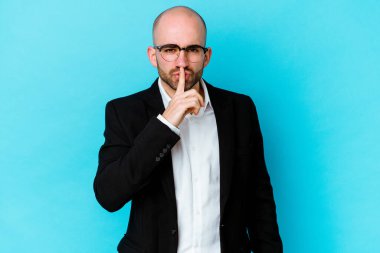 Genç beyaz tenli kel adam mavi arka planda izole edilmiş bir şekilde bir sır saklıyor ya da sessizlik istiyor..