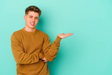 Mavi arka planda izole edilmiş genç beyaz bir adam bir palmiye üzerinde fotokopi alanı ve belinde bir el daha gösteriyor..