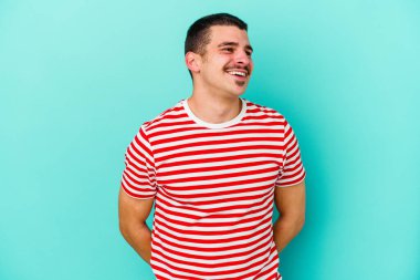 Kafkasyalı genç adam mavi arka planda izole edilmiş rahat ve mutlu bir şekilde gülüyor, boynunda dişlerini gösteriyor..