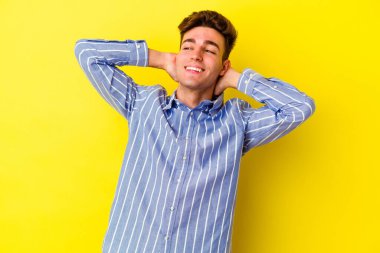 Sarı arka planda izole edilmiş genç beyaz bir adam kendini güvende hissediyor, elleri başının arkasında..