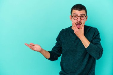 Mavi arka planda izole edilmiş genç beyaz bir adam avucunun içinde fotokopi alanı tutuyor, el üstünde tutuyor. Şaşırmış ve mutlu..