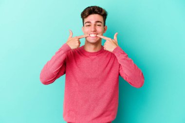 Mavi arka planda izole edilmiş genç beyaz adam gülümsüyor, parmaklarını ağıza doğrultuyor..