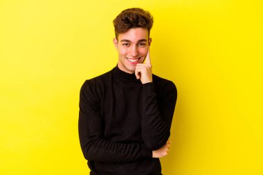 Sarı arka planda izole edilmiş genç, mutlu ve kendinden emin gülümseyen, elleriyle çenesine dokunan beyaz bir adam..