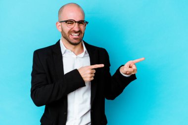 Genç, beyaz, kel bir adam mavi arka planda izole edilmiş ve işaret parmaklarıyla fotokopi alanını işaret ediyor. Heyecan ve arzuyu ifade ediyor..
