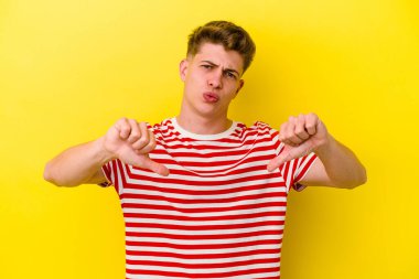 Sarı arka planda izole edilmiş genç beyaz adam baş parmağını aşağı gösteriyor ve hoşlanmadığını ifade ediyor..