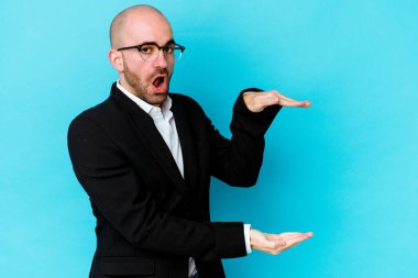 Genç, beyaz, kel bir adam mavi arka planda izole edilmiş. Elleri arasında bir kopya alanı tutarken şok olmuş ve hayrete düşmüştü..