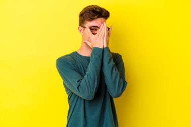 Sarı arka planda izole edilmiş genç beyaz adam kameraya parmaklarıyla göz kırpıyor, yüzünü kapatmaktan utanıyor..