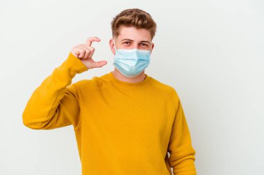 Koronavirüs için maske takan genç bir adam beyaz arka planda izole edilmiş küçük bir şey tutuyor, gülümsüyor ve kendinden emin..