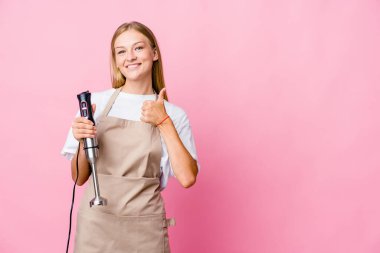 Genç Rus aşçı kadın, elinde elektrikli mikser tutarken izole edilmiş gülümsüyor ve başparmağını kaldırıyor.