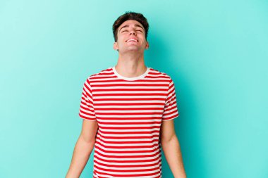 Kafkasyalı genç adam mavi arka planda izole edilmiş rahat ve mutlu bir şekilde gülüyor, boynunda dişlerini gösteriyor..