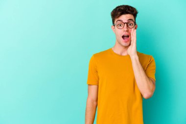 Mavi arka planda izole edilmiş genç beyaz adam yüksek sesle bağırıyor, gözleri açık ve elleri gergin tutuyor..