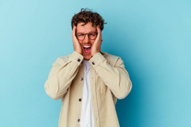 Gözlüklü genç beyaz adam mavi arka planda izole edilmiş kulakları elleriyle kapatıyor ve çok fazla ses duymamaya çalışıyor..