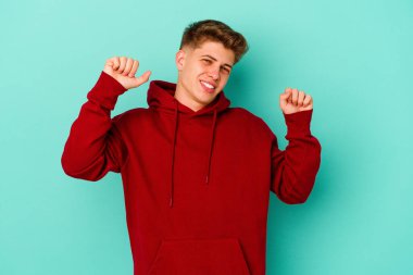 Mavi arka planda izole genç beyaz adam özel bir günü kutluyor, atlıyor ve enerjiyle kollarını kaldırıyor..