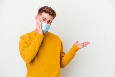 Koronavirüs için maske takan genç bir adam beyaz arka planda izole edilmiş bir şekilde bir avuç içinde fotokopi alanı tutuyor, el üstünde tutuyor. Şaşırmış ve mutlu..