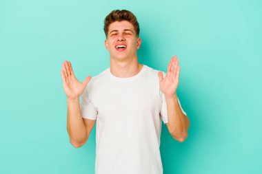 Mavi arka planda izole olmuş genç beyaz adam yüksek sesle gülüyor ve elini göğsüne koyuyor..