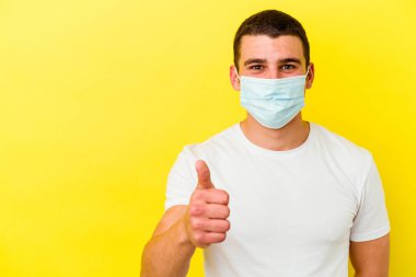 Koronavirüs için koruyucu takan genç beyaz adam sarı arka planda izole edilmiş gülümsüyor ve başparmağını kaldırıyor.