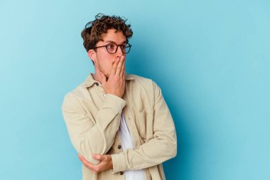 Mavi arka planda izole edilmiş gözlük takan genç beyaz bir adam esneyerek ağzını eliyle kapatan yorgun bir jest yapıyor..