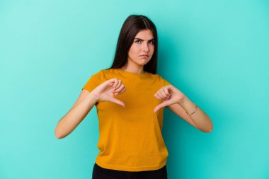 Mavi arka planda izole edilmiş genç beyaz kadın baş parmağını aşağı gösteriyor, hayal kırıklığı konsepti.