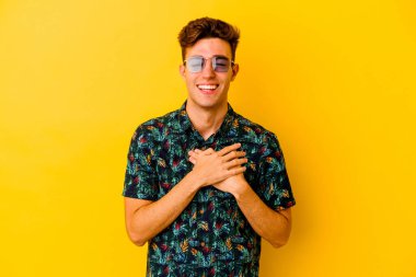 Sarı arka planda izole edilmiş Hawaii tişörtü giyen genç beyaz adam gülüyor, elleri yüreğinde, mutluluk kavramında..