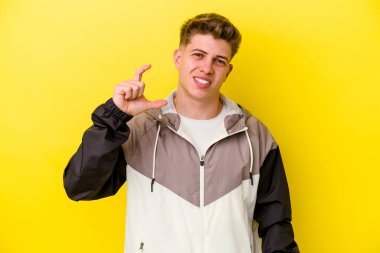 Sarı arka planda izole edilmiş genç beyaz adam işaret parmaklarıyla küçük bir şey tutuyor, gülümsüyor ve kendinden emin..