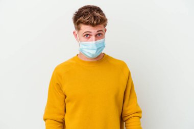 Koronavirüs maskesi takan genç adam beyaz arka planda izole edilmiş omuzlarını silker ve gözleri açık kalır..