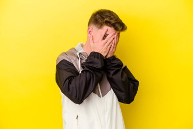 Sarı arka planda izole edilmiş genç beyaz adam parmaklarının arasından korkmuş ve gergin bir şekilde göz kırp..