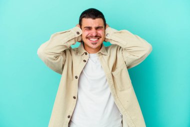 Kafkasyalı genç adam mavi arka planda izole edilmiş kulakları kulaklarıyla kapatıyor çok fazla ses duymamaya çalışıyor..