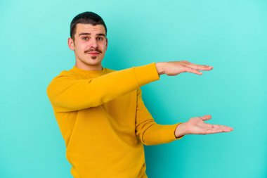 Mavi arka planda izole genç beyaz adam iki eliyle bir şey tutuyor, ürün sunumu..