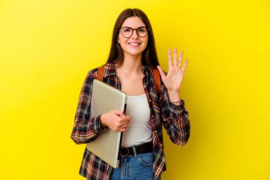 Pembe arka planda dizüstü bilgisayar tutan beyaz, genç bir bayan beş numarayı parmaklarıyla gülümsüyor..