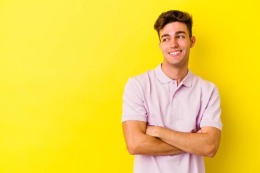 Sarı arka planda izole edilmiş genç beyaz adam kollarını kavuşturarak gülümsüyor..