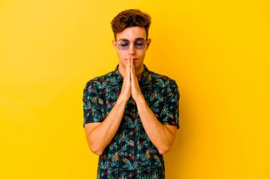 Sarı arka planda izole edilmiş Hawaii tişörtü giyen genç beyaz bir adam el ele tutuşup ağzının yanında dua ediyor, kendinden emin hissediyor..