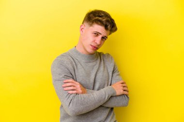 Sarı arka planda izole edilmiş genç beyaz adam hoşnutsuzca kaşlarını çatmış, kollarını kavuşturmuş..