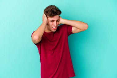 Mavi arka planda izole edilmiş genç beyaz adam kulaklarını elleriyle kapatıyor..