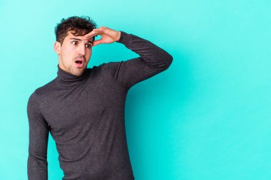 Kafkasyalı genç adam mavi arka planda izole edilmiş, uzaklara bakıyor ve elini alnına koyuyor..