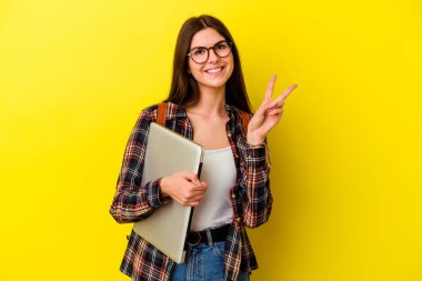 Pembe arka planda dizüstü bilgisayarı olan beyaz bir kadın öğrenci neşeli ve kaygısız parmaklarıyla barış sembolü gösteriyor..