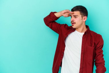 Kafkasyalı genç adam mavi arka planda izole edilmiş, uzaklara bakıyor ve elini alnına koyuyor..