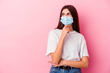 Pembe arka planda izole edilmiş virüs maskesi takan beyaz bir kadın şüpheli ve şüpheci bir ifadeyle yana bakıyor..