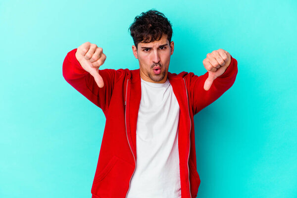 Молодой кавказский мужчина на голубом фоне показывает большой палец вниз и выражает неприязнь.