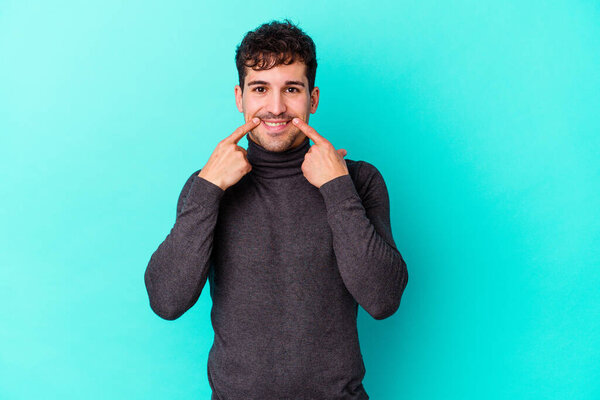Молодой кавказский мужчина на голубом фоне улыбается, указывая пальцем на рот.