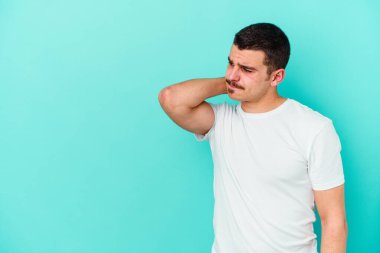 Kafkasyalı genç adam mavi arka planda izole edilmiş kafasının arkasına dokunuyor, düşünüyor ve bir seçim yapıyor..