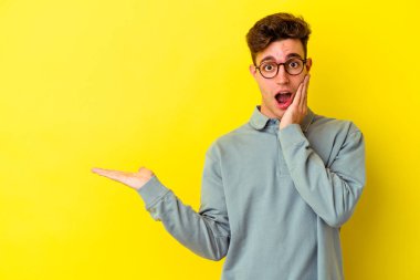 Sarı arka planda izole edilmiş genç beyaz adam, avuç içi fotokopi alanını elinde tutmaktan etkilendi..