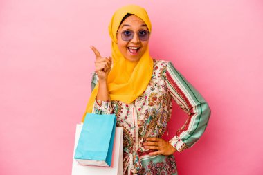 Genç Müslüman kadın pembe arka planda izole giysiler alıyor. Bir fikri, ilham verici bir konsepti var..