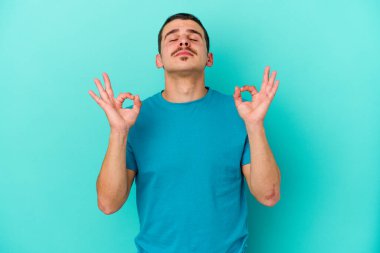 Mavi arka planda izole edilmiş genç beyaz adam yorucu bir günün ardından rahatlıyor. Yoga yapıyor..