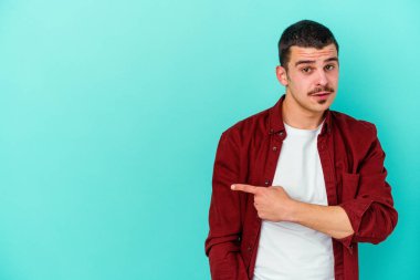 Mavi arka planda izole edilmiş genç beyaz adam gülümsüyor ve boş alanda bir şeyler gösteriyor..