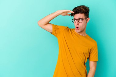 Kafkasyalı genç adam mavi arka planda izole edilmiş, uzaklara bakıyor ve elini alnına koyuyor..