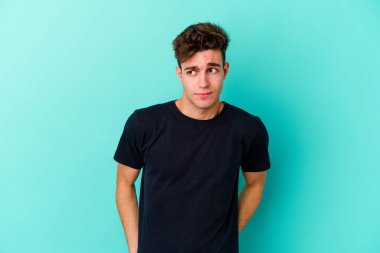 Kafkasyalı genç adam mavi arka planda izole, kafası karışmış, şüpheli ve güvensiz hissediyor..