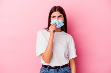 Pembe arka planda izole edilmiş, tırnak yiyen, gergin ve endişeli virüs maskesi takan beyaz bir kadın..